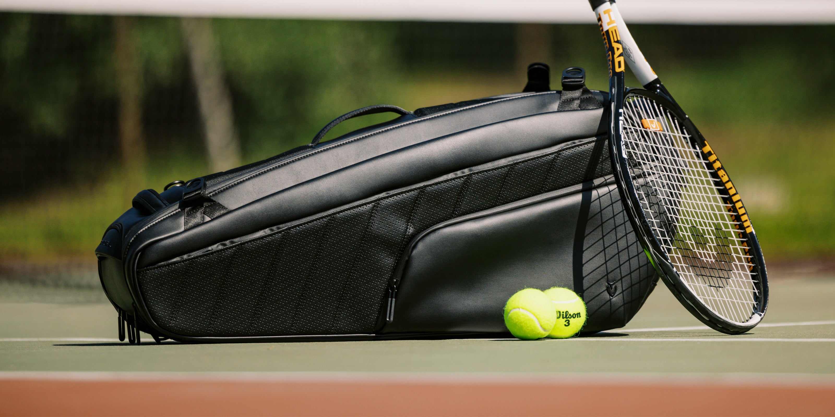 Leather black racquet bag on tennis court with 2 tennis balls and a tennis racquet