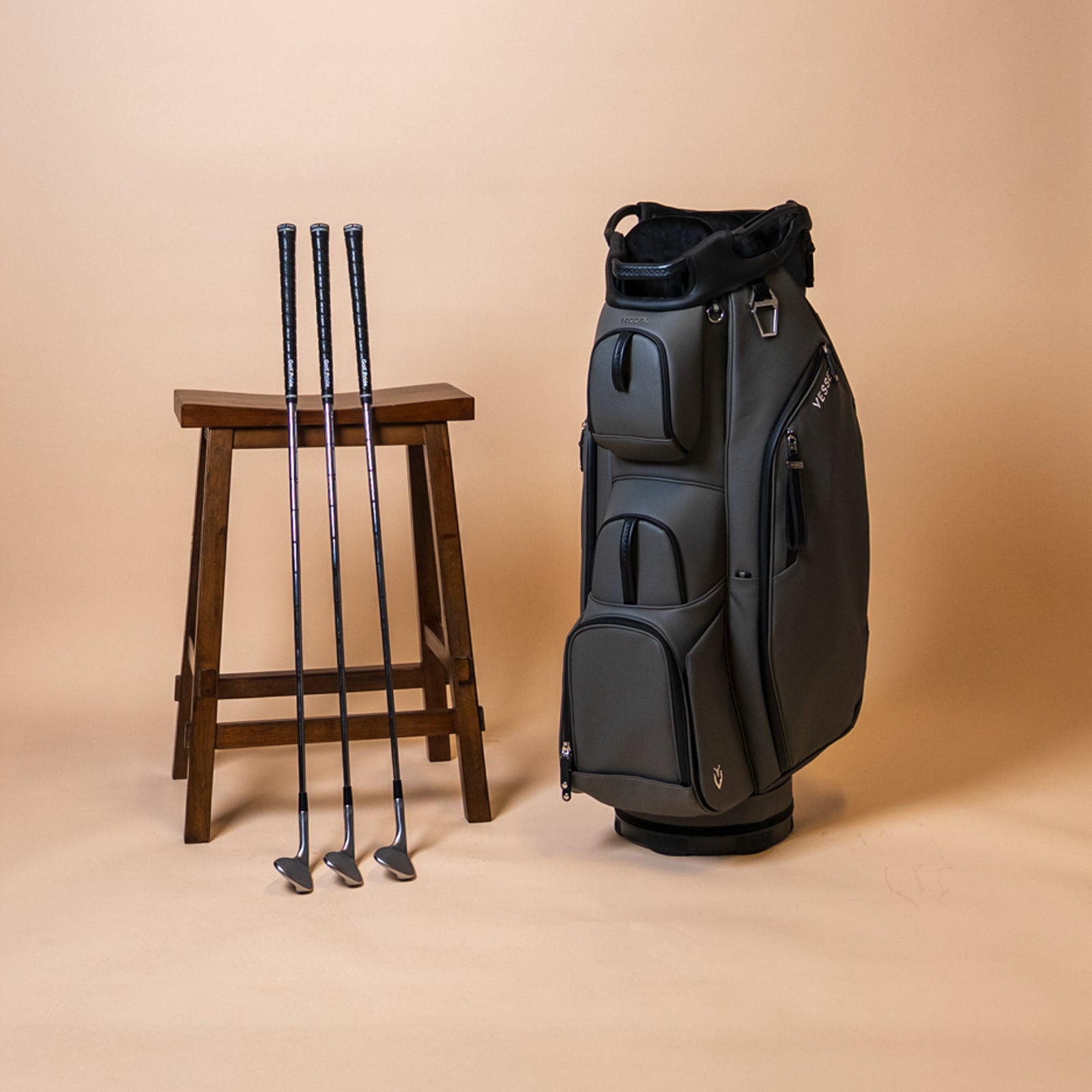 Grey cart bag on a taupe background next to a stool and three golf clubs