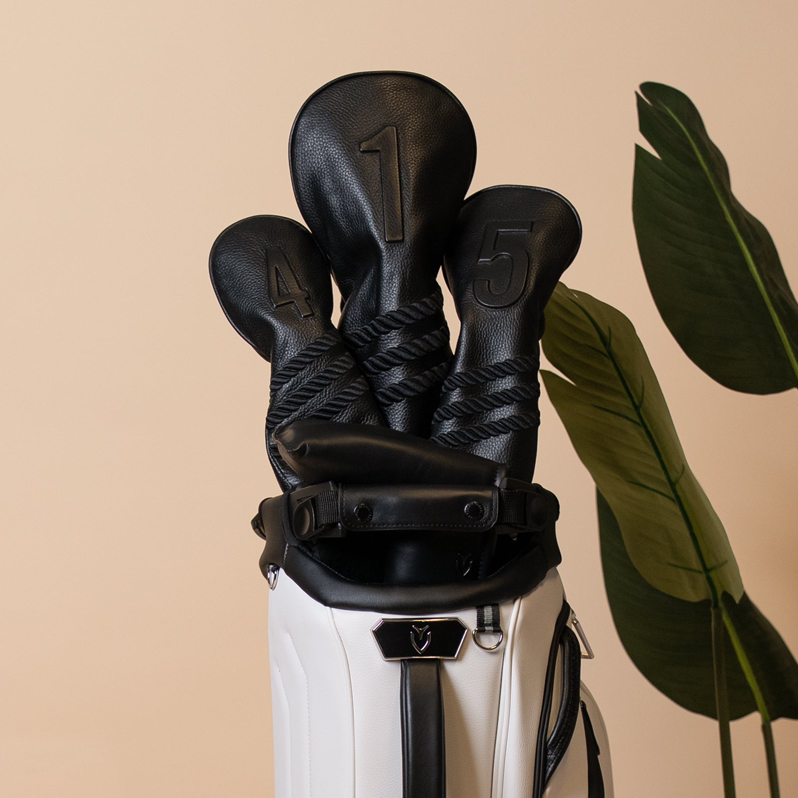 Black leather headcovers in a white leather golf bag next to plant