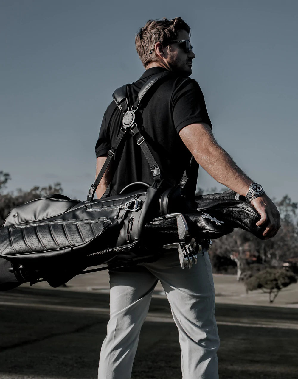 A man wearing a black shirt and sunglasses carries a black golf bag filled with golf clubs on his back outside