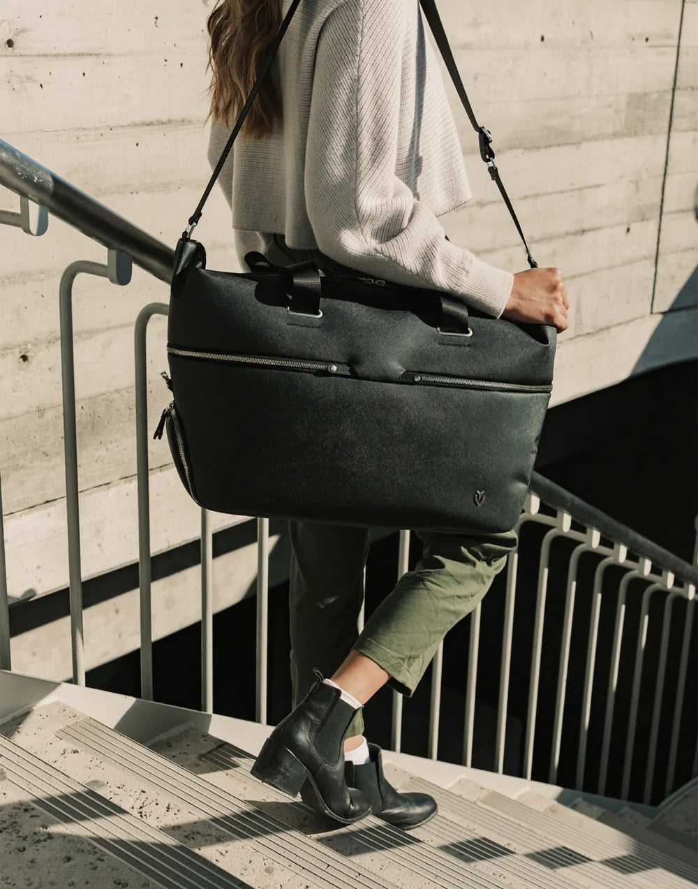 Woman wearing a beige sweater carries a black leather duffel bag as she walks down the stairs