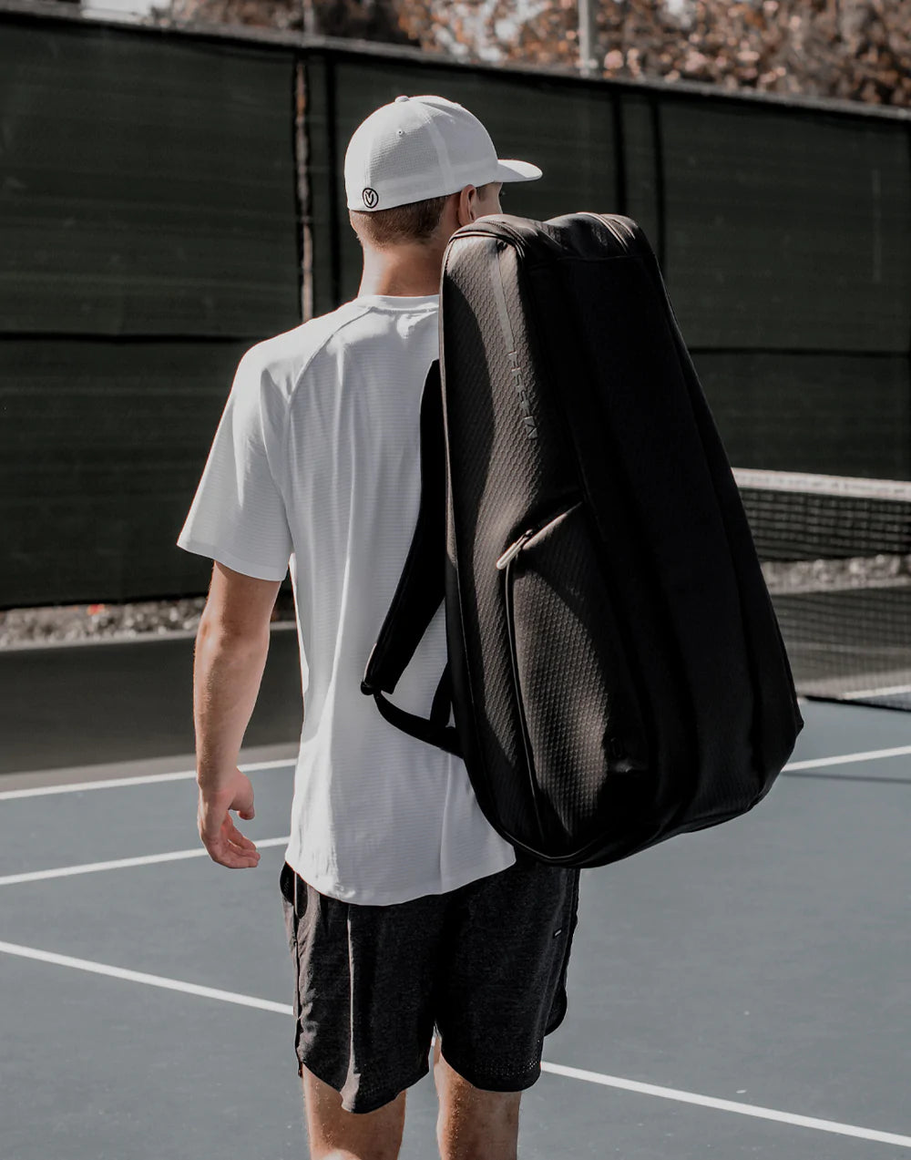 A man wearing a white hat and shirt carries a black tennis bag on his back on the tennis court