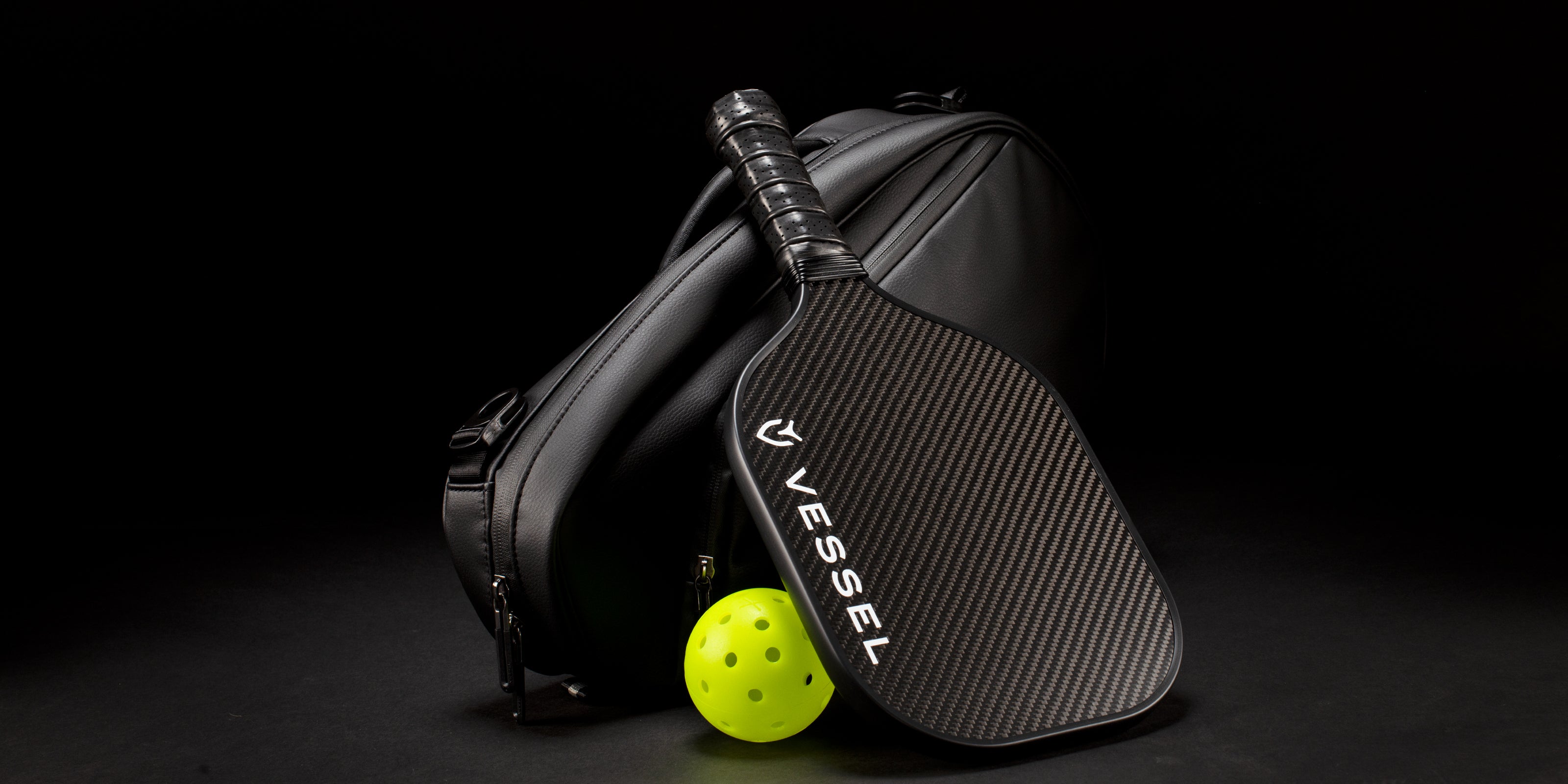 A black Carbon Pickleball Paddle rests against a black Pickleball Lite Paddle Bag along with a yellow pickleball on a black background