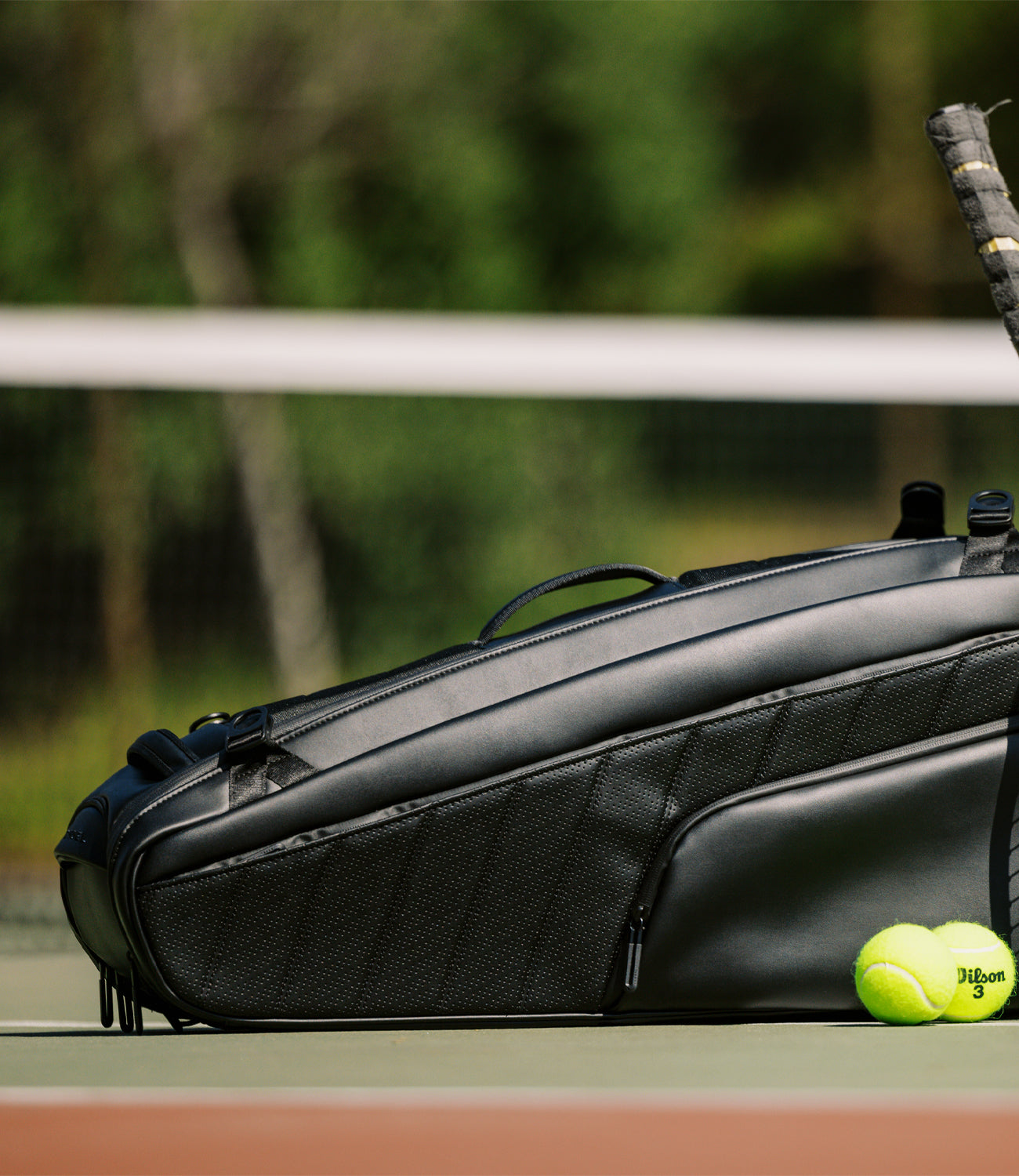 Leather black racquet bag on tennis court with 2 tennis balls and a tennis racquet