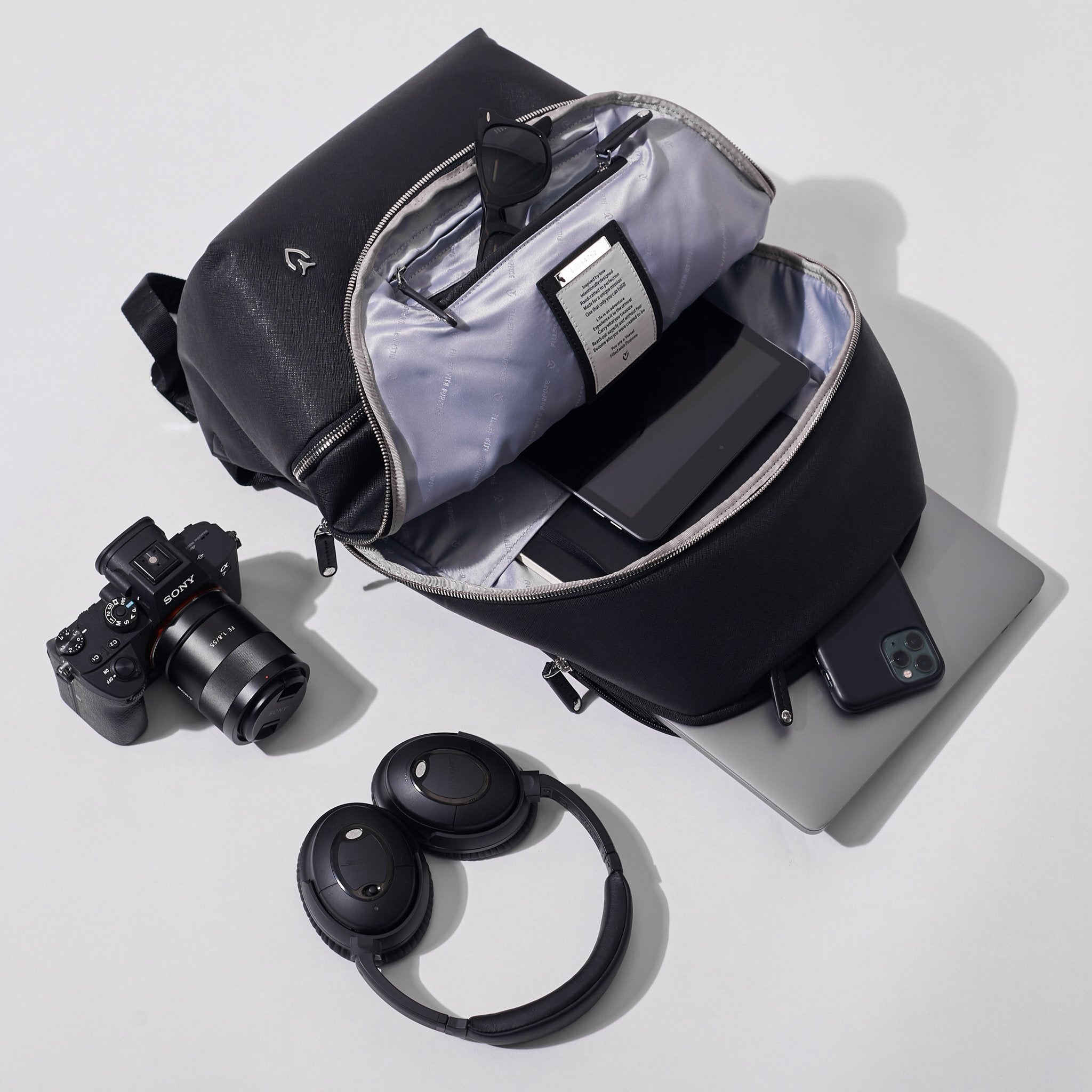 A crosshatch black signature backpack on a white background. A computer, phone. camera, headphones surround the bag