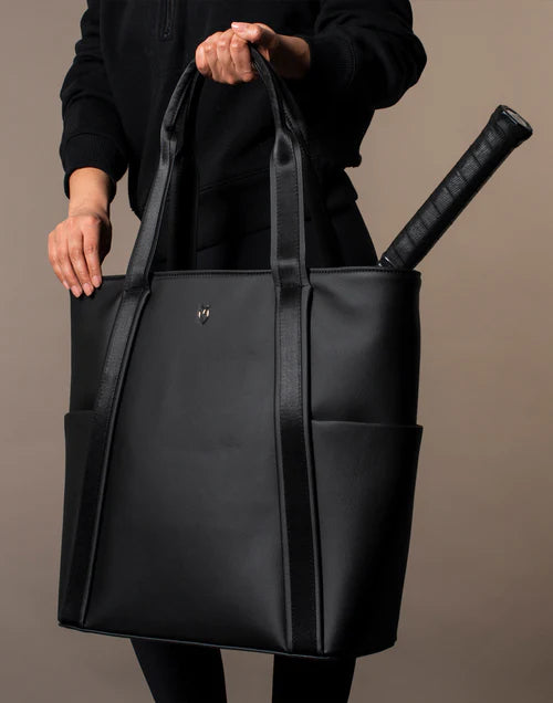 Model wearing all black holds the black Baseline Tennis tote in a brown studio