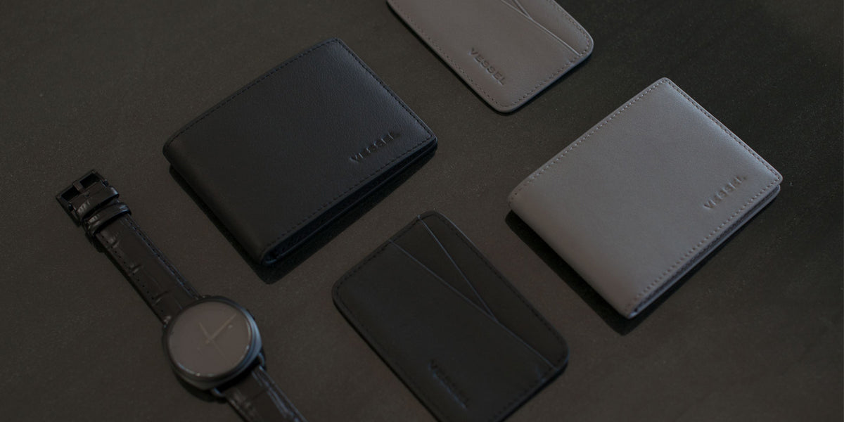 A watch is placed next to two Leather Bifold Wallets and one Leather Cardholder wallet on a brown background