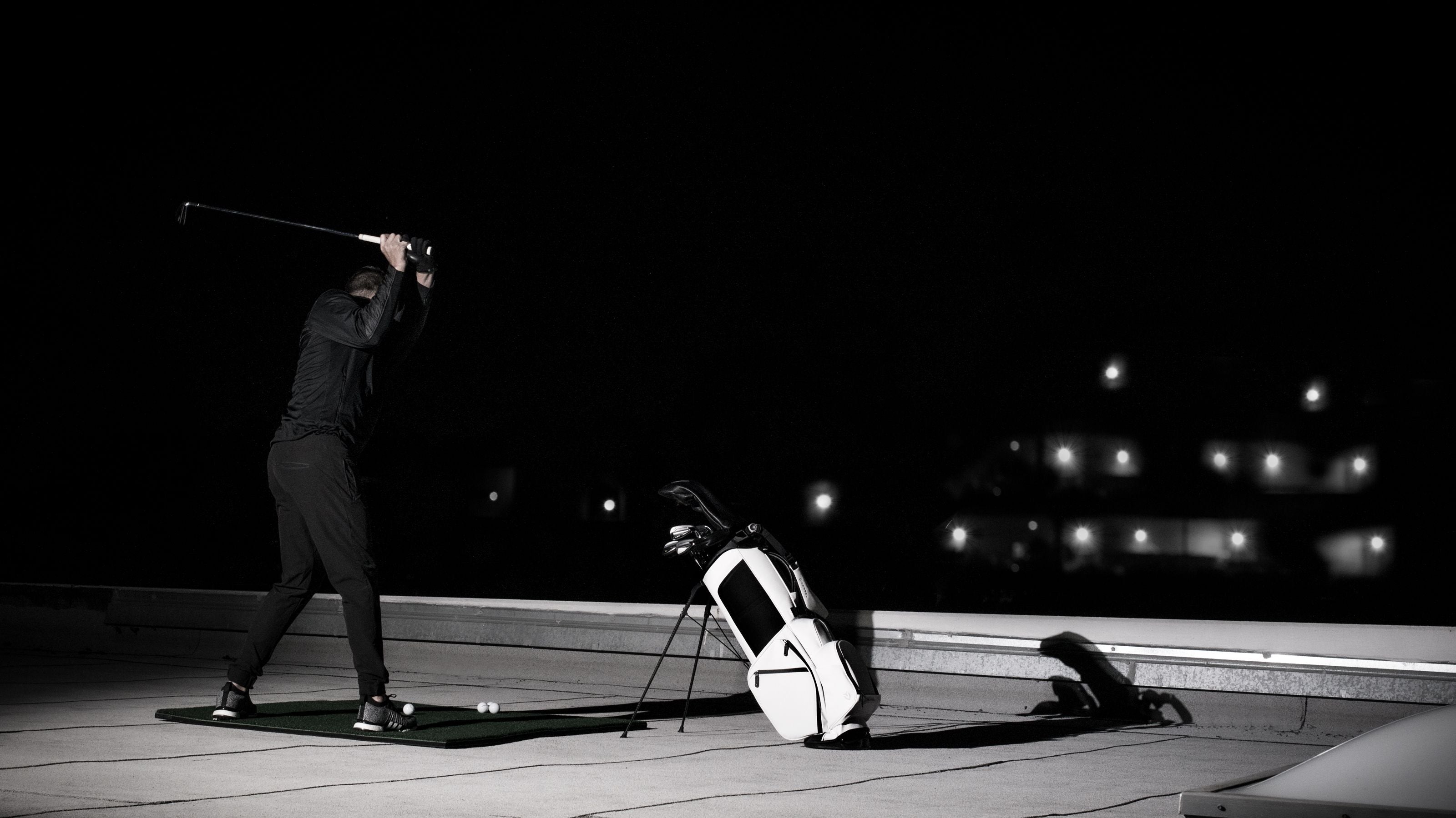 White golf bag is propped up on the ground next to a person playing golf on a roof at night