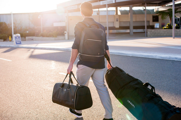 A man is walking into an airport with a golf travel bag and garment duffel