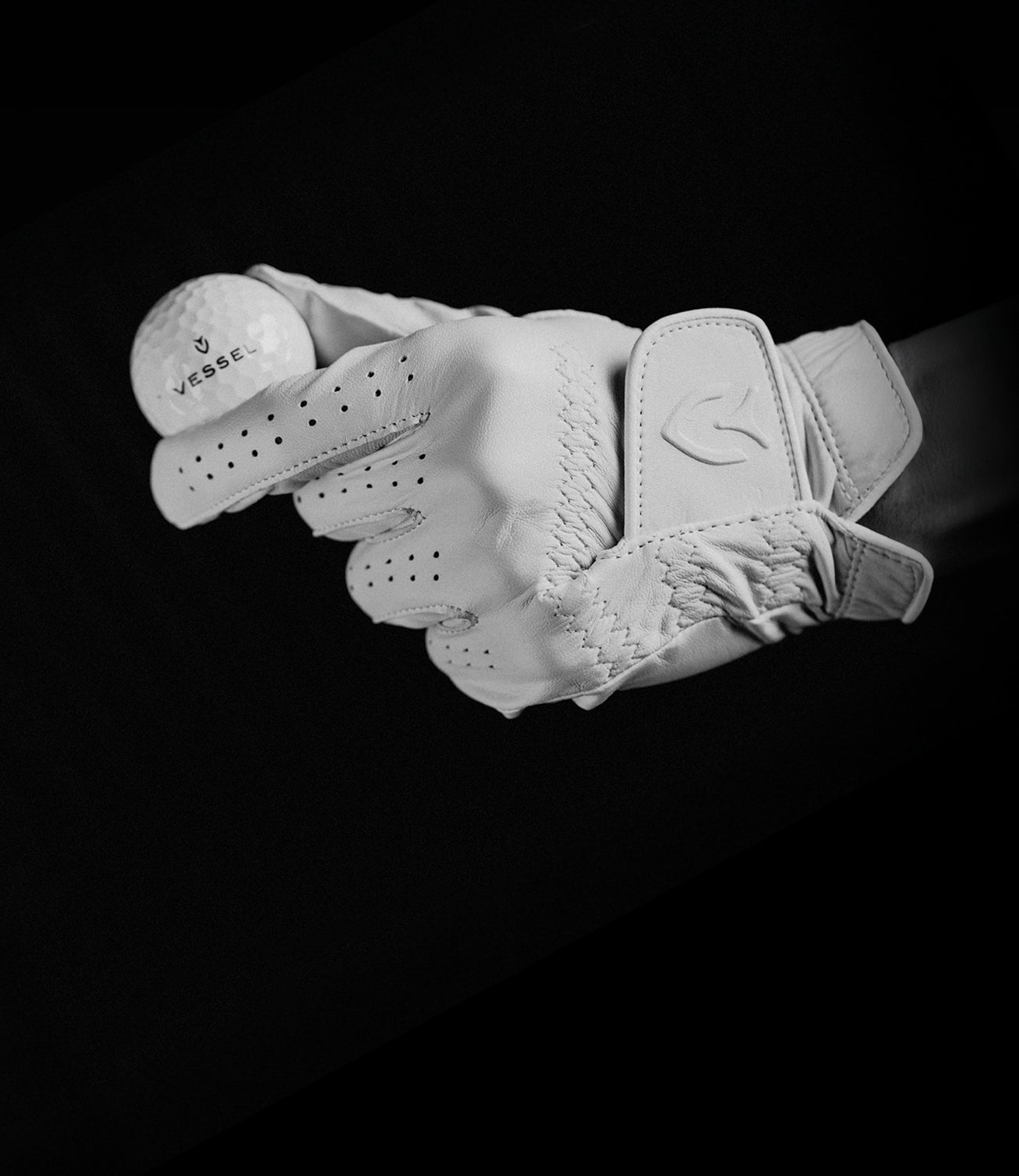 Person holding a golf ball marked with a Vessel logo, while wearing a white leather golf glove