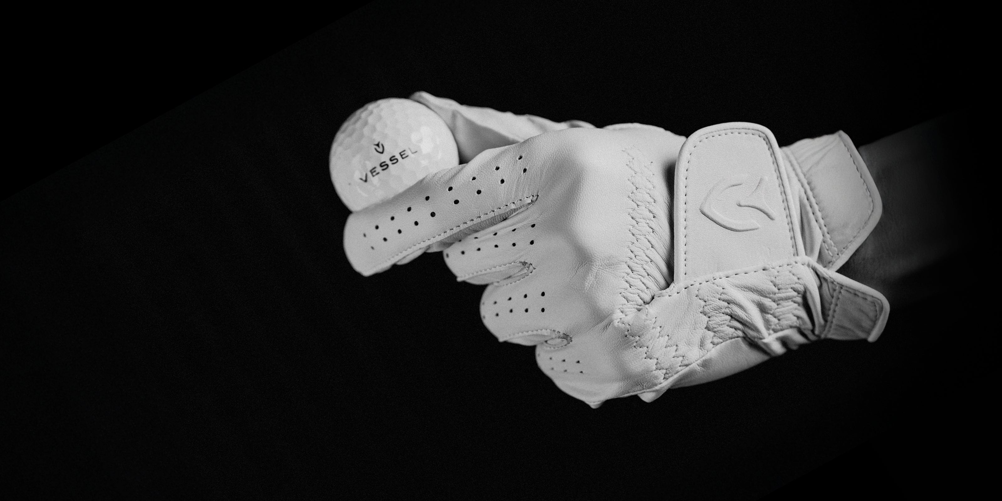 Person holding a golf ball marked with a Vessel logo, while wearing a white leather golf glove