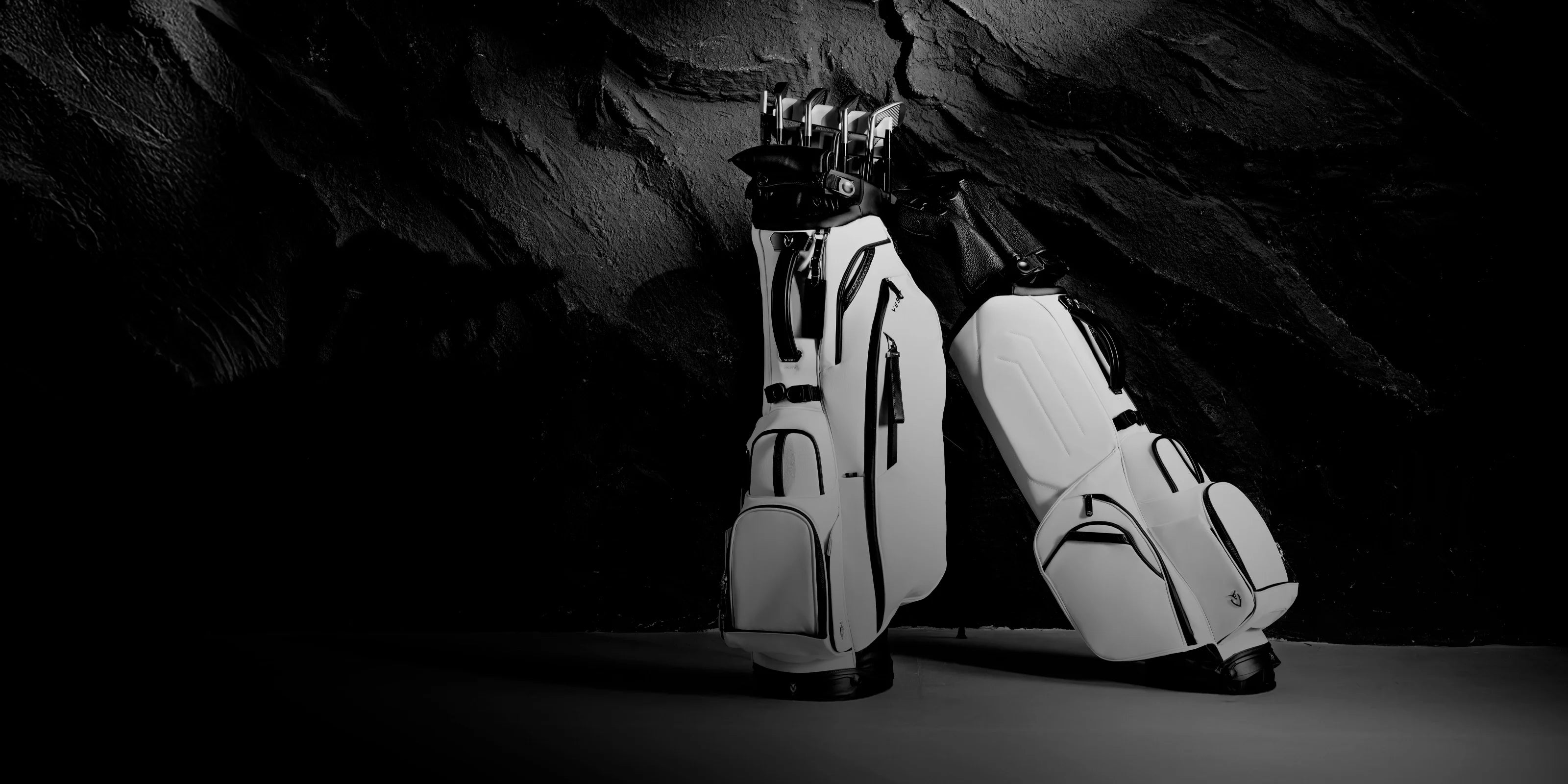 Two white leather golf stand bags against a dark, rocky wall