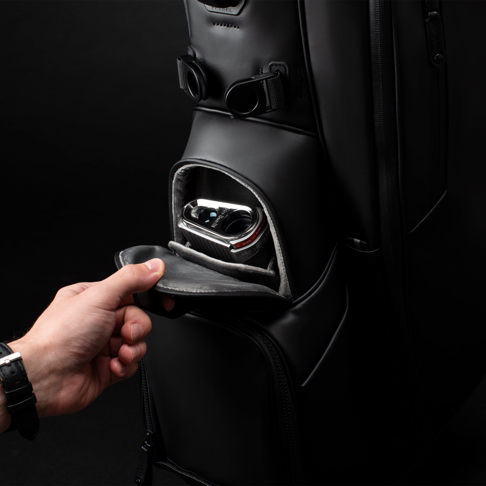 Close up image of a hand opening the magnetic pocket on a black golf bag in a black studio