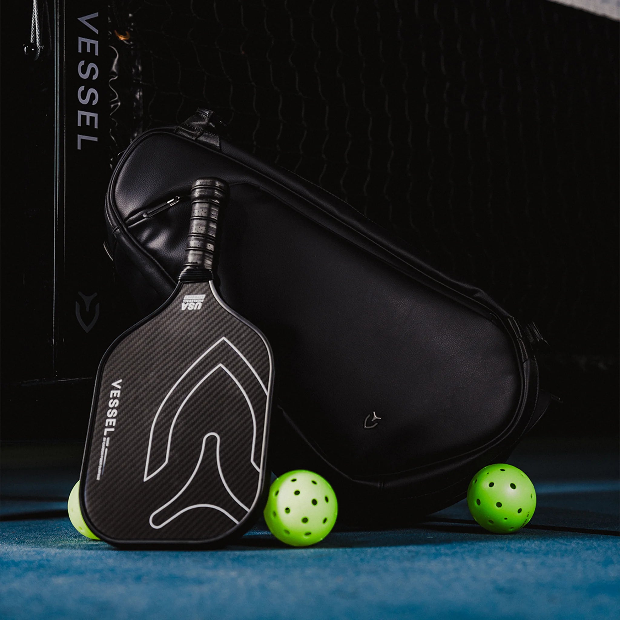 Black carbon fiber pickleball paddle leaning against Vessel black leather pickleball paddle bag with three pickleballs on the ground