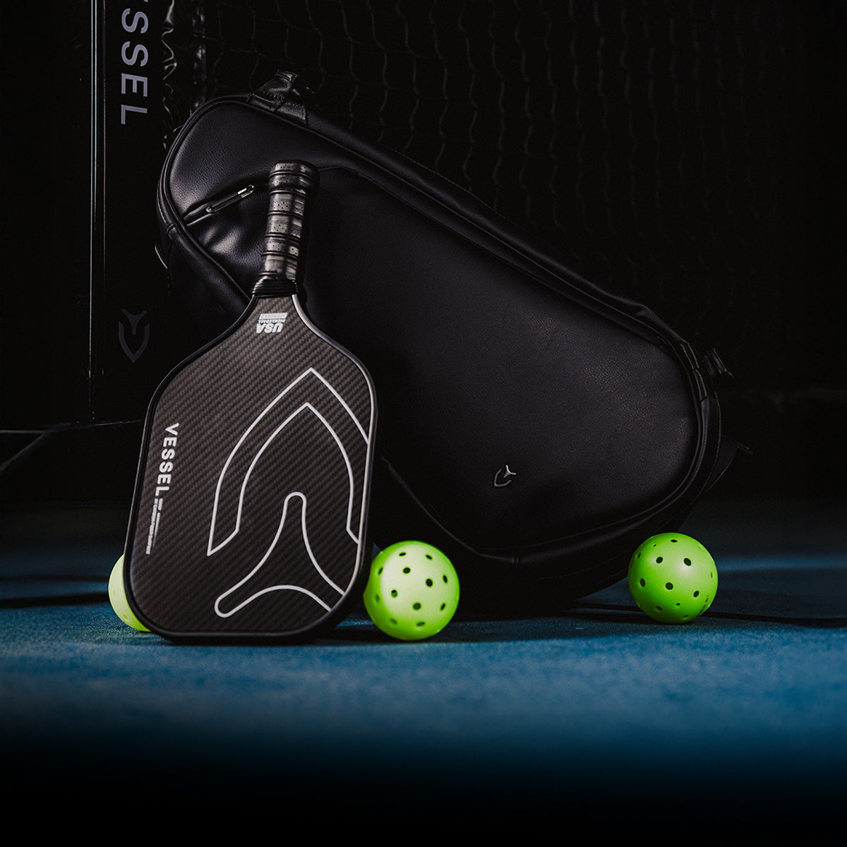 Carbon pickleball paddle leaning against a black leather pickleball paddle bag with two pickleballs on the ground 