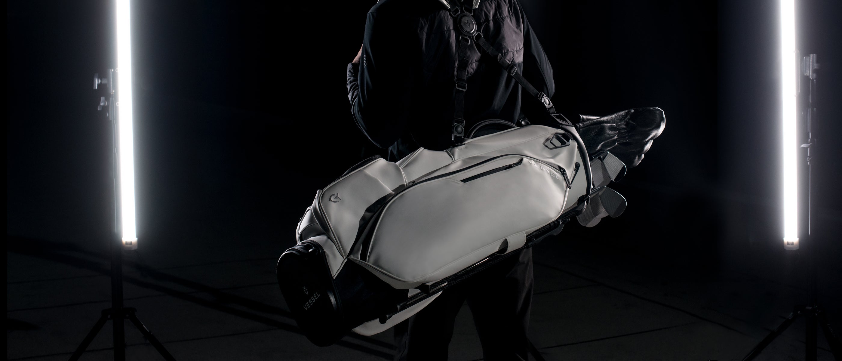 Model in black carries a white golf stand bag on his back in a dark studio with two studio lights