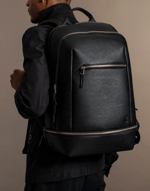 Model wearing all black carries a black leather backpack in a brown studio