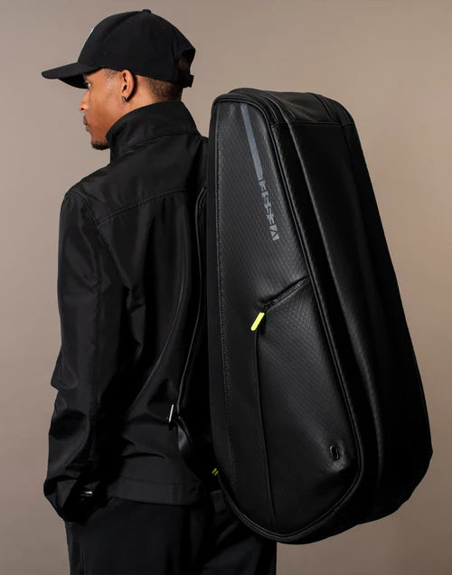 Man wearing black and a black hat carries the black Baseline Raquet Bag on his back in a brown studio