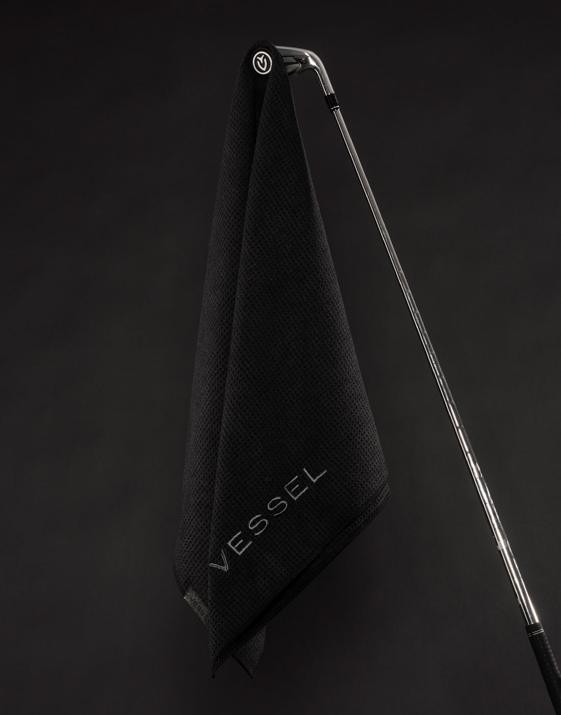 A black golf towel hangs from a silver golf club in a black studio